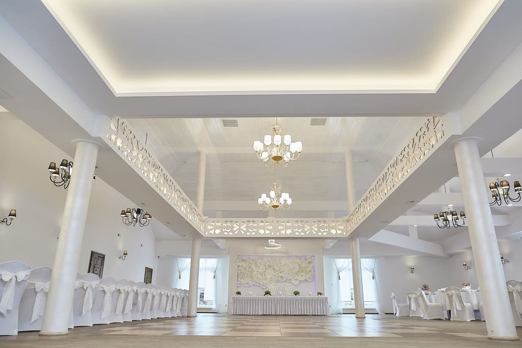 Duża sala balowa z białymi kolumnami, ażurowymi zdobieniami i żyrandolami